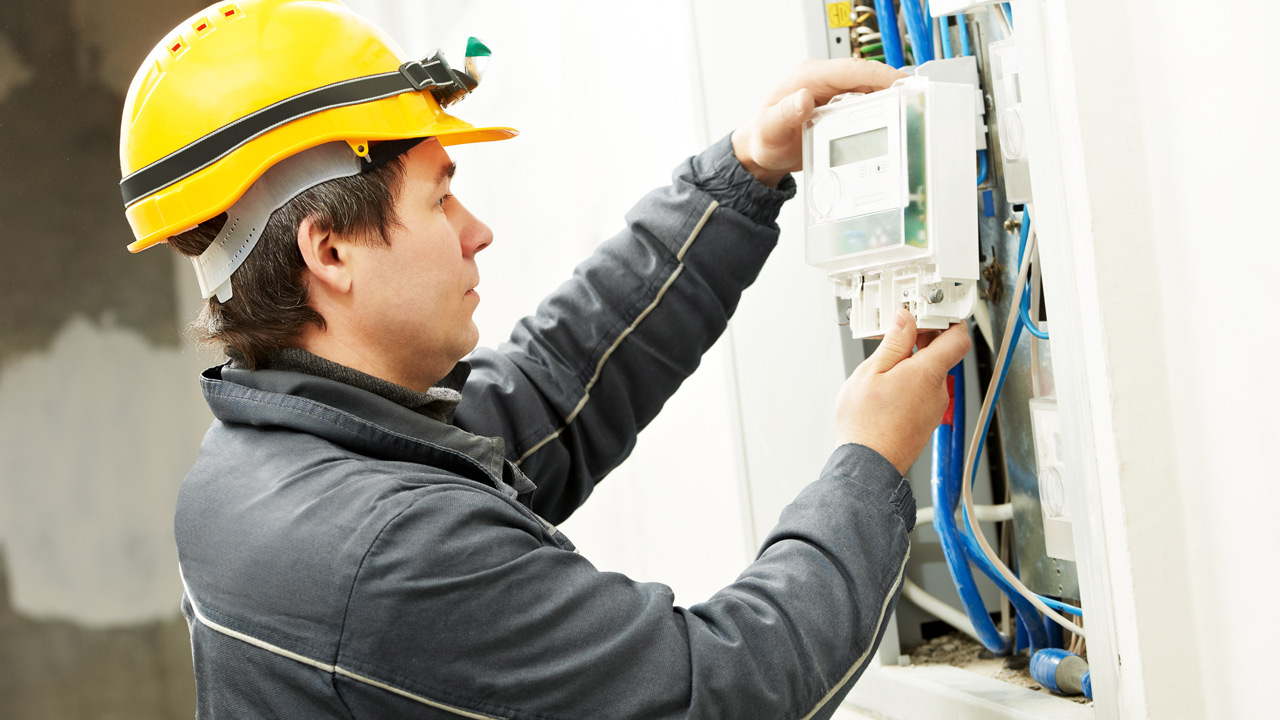 ifi_ Ein Arbeiter, der einen gelben Schutzhelm und eine graue Jacke trägt, installiert oder inspiziert eine elektrische Schalttafel an einer Wand, die von blauen und weißen Kabeln umgeben ist.