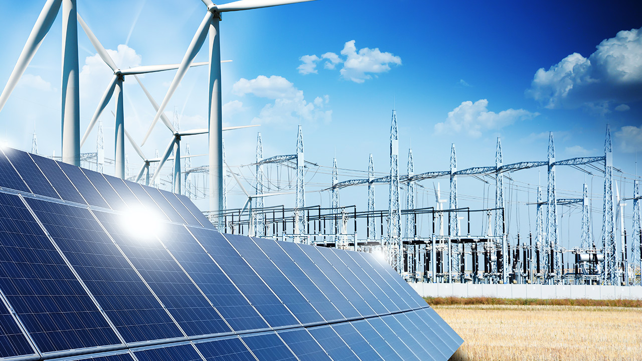 ifi_ Im Vordergrund sind Reihen von Sonnenkollektoren und Windturbinen zu sehen, im Hintergrund ein Umspannwerk und Stromleitungen unter einem strahlend blauen Himmel mit vereinzelten Wolken.