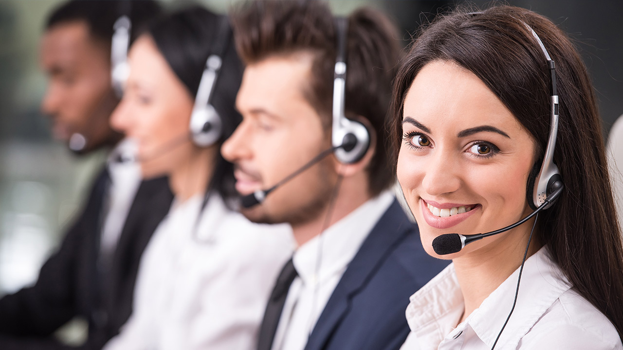 ifi_ Vier Kundendienstmitarbeiter, die Headsets tragen, sitzen in einer Reihe. Die Frau im Vordergrund lächelt in die Kamera, während die anderen auf ihre Arbeit konzentriert sind.