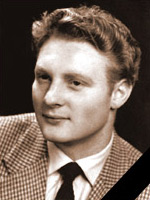 ifi_ Ein junger Mann mit hellem Haar, der eine karierte Anzugsjacke, ein weißes Hemd und eine dunkle Krawatte trägt, posiert für ein formelles Porträt vor einem dunklen Hintergrund.