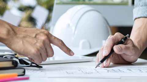 ifi_ Zwei Personen begutachten Architekturpläne auf einem Schreibtisch; eine zeigt auf etwas, während die andere einen Stift hält. Im Hintergrund sind ein weißer Schutzhelm und Papiere zu sehen.